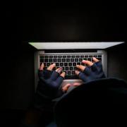 An anonymous hooded figure sitting in the dark typing on a laptop. Credit: Canva